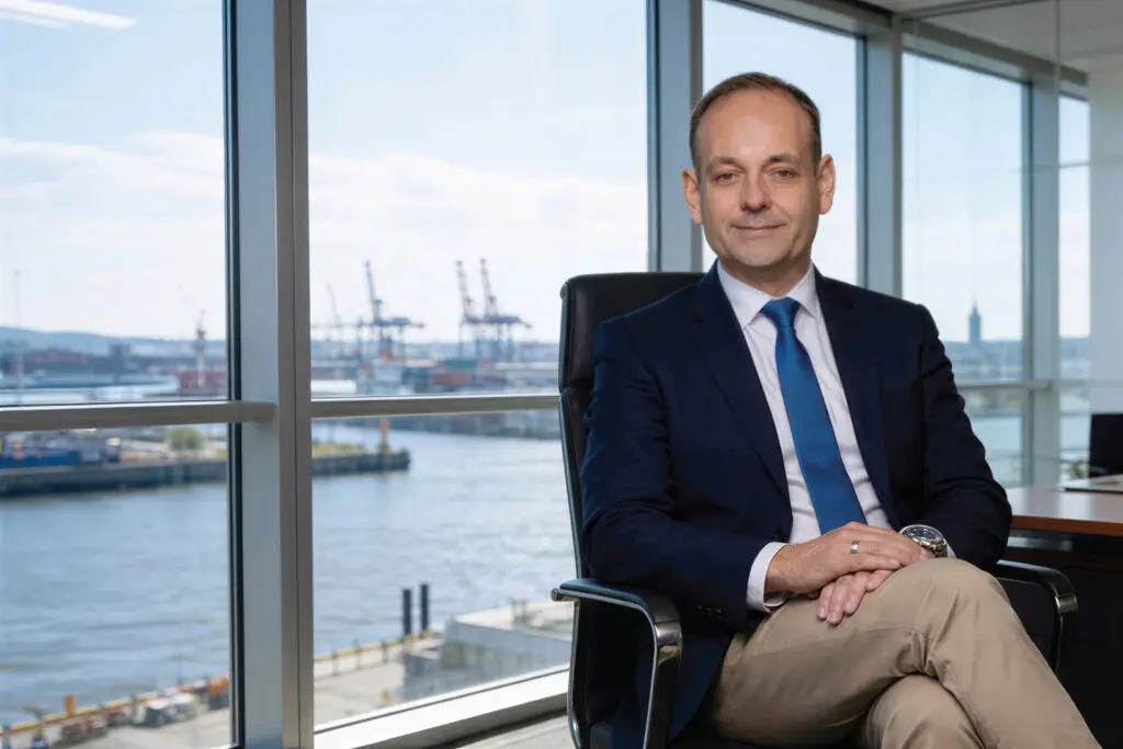 Portrait von Jens Wolfhagen sitzt im Büro mit Blick auf Hafenkräne und Elbe im Hintergrund.
